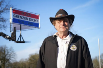 Holenderski aktywista Jan Huzen w czarnym kapeluszu na tle transparentu z hasłem o zamknięciu granic, skazany za planowane obywatelskie kontrole graniczne.